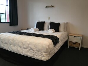 A neatly made bed with white and black bedding, a bedside table with a basket, and a window with curtains. - Kiwi Court Motel (Hawera)