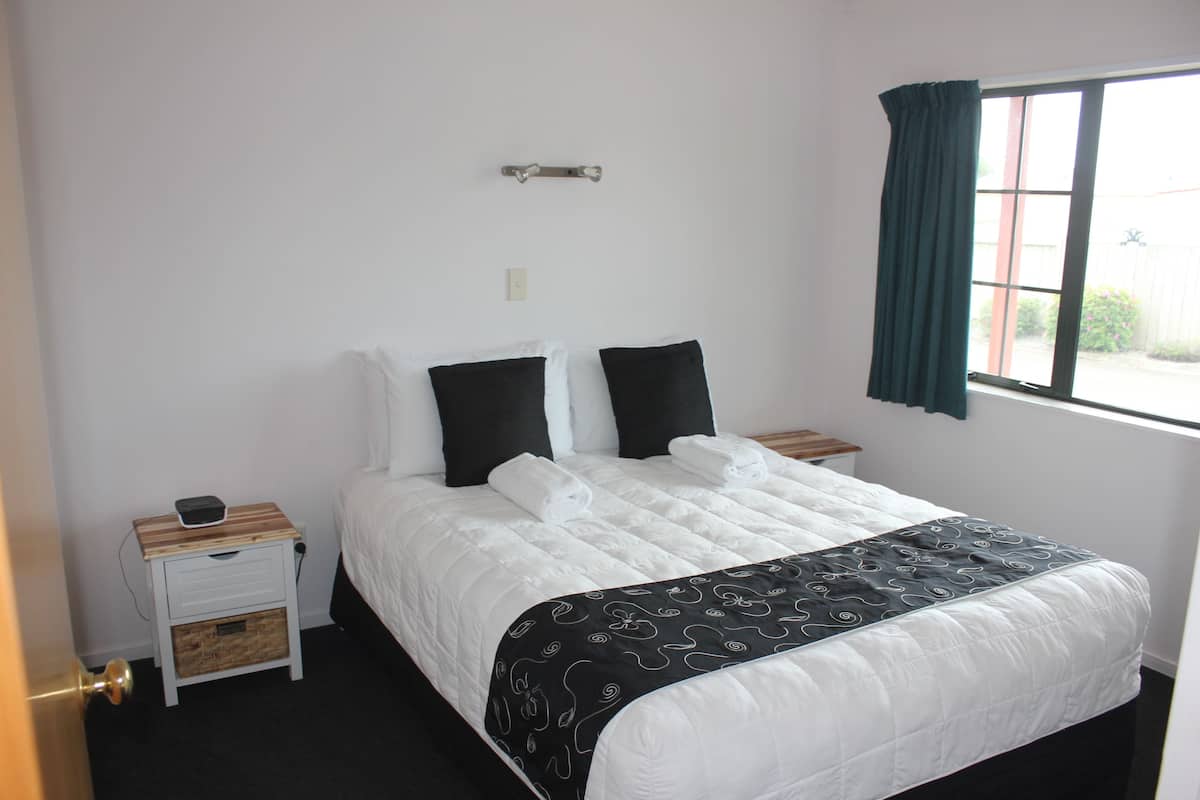 A neatly made bed with white bedding and a black and white patterned throw, a small bedside table with a lamp, and a window with a green curtain.