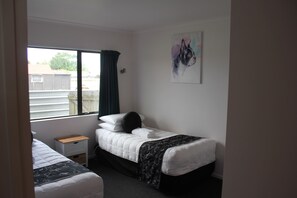 A bedroom with two single beds, a small bedside table, a window with curtains, and a wall-mounted painting. - Kiwi Court Motel (Hawera)