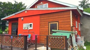 Ett rött trähus med en liten veranda, ett grönt solskydd och en inhägnad uteplats.