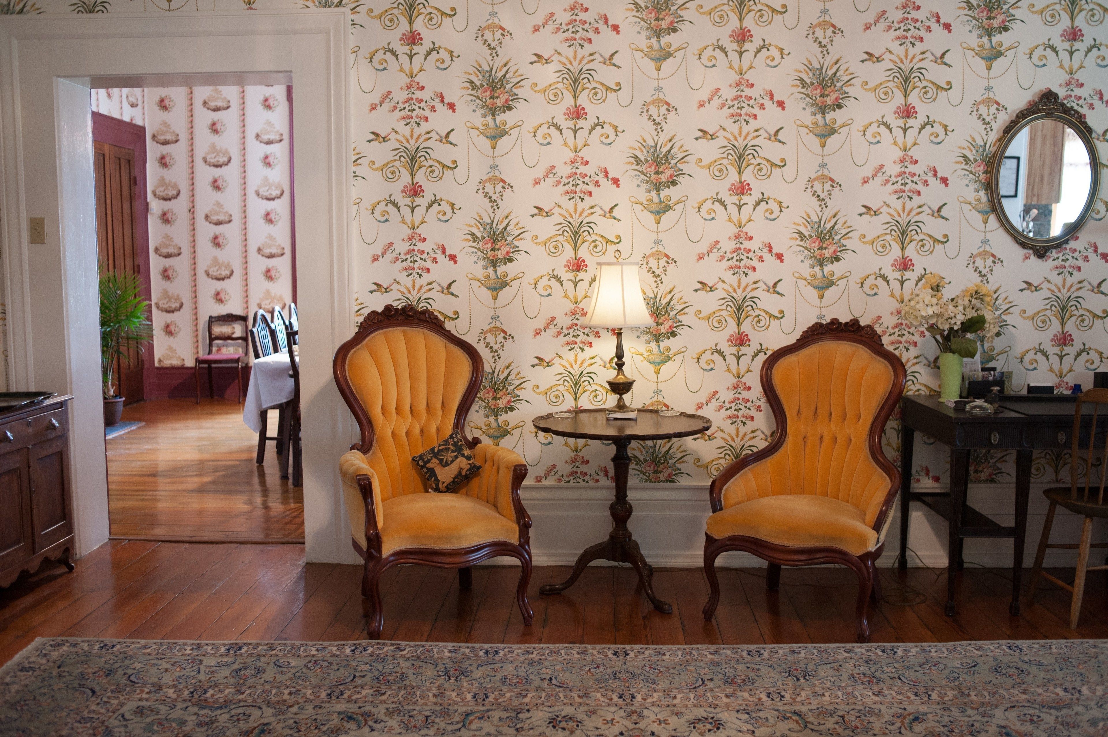 A room with two upholstered chairs, a small round table with a lamp, and floral wallpaper.