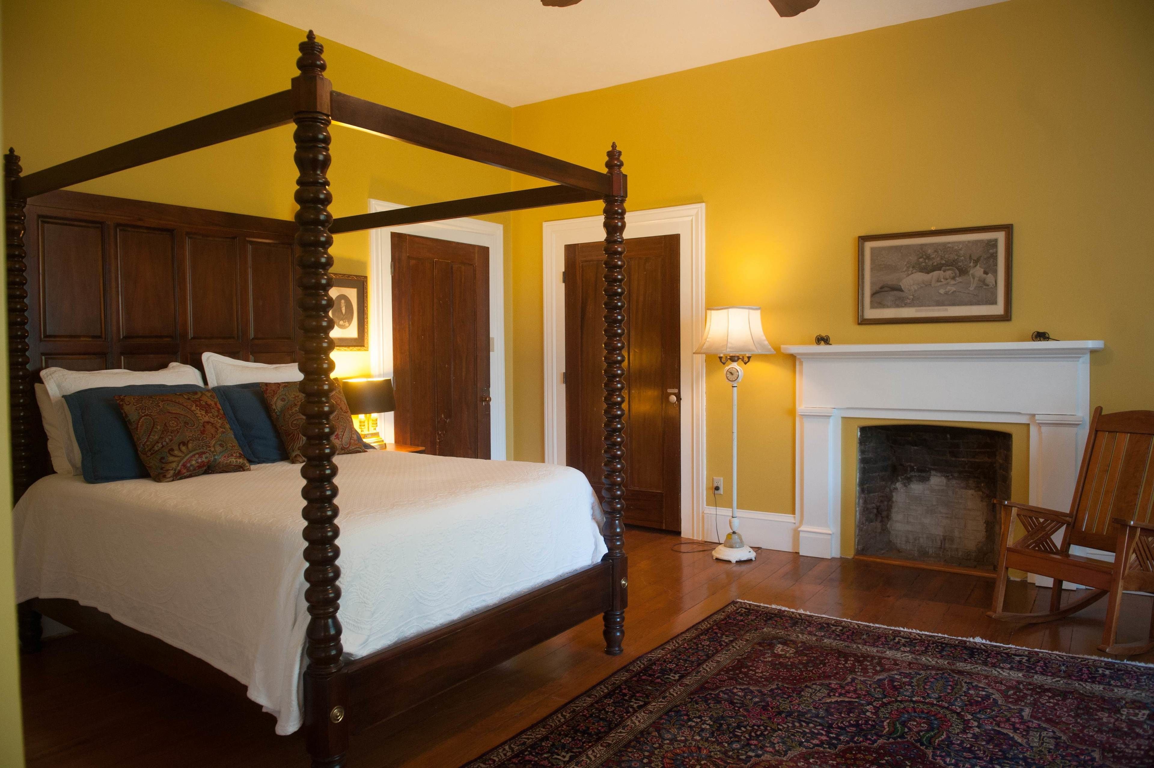 A four-poster bed with a white bedspread and blue pillows, a wooden floor, a fireplace with a mantel, and a framed picture above it.