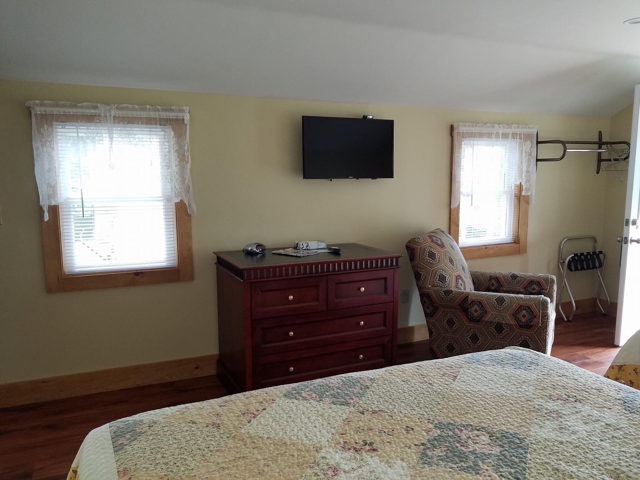A bedroom with a bed, a dresser, a chair, a TV, and two windows with curtains.