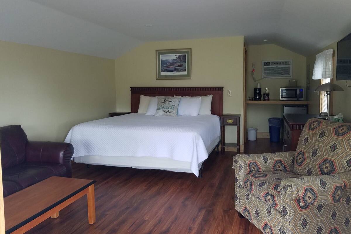 A hotel room with a large bed, a wooden headboard, a brown leather chair, a patterned sofa, a small table, a desk with a computer, and a microwave.