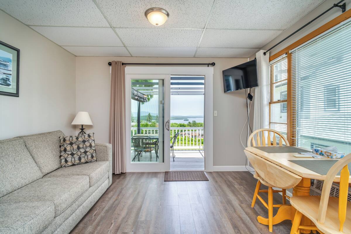 A room with a sofa, a table with chairs, a TV mounted on the wall, and a sliding glass door leading to an outdoor area.