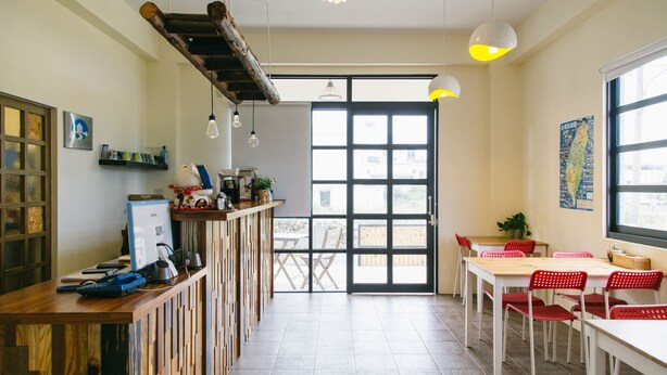 Una cafetería moderna con mostradores de madera, mesas blancas y sillas rojas. Se observa un gran ventanal con vistas al exterior.