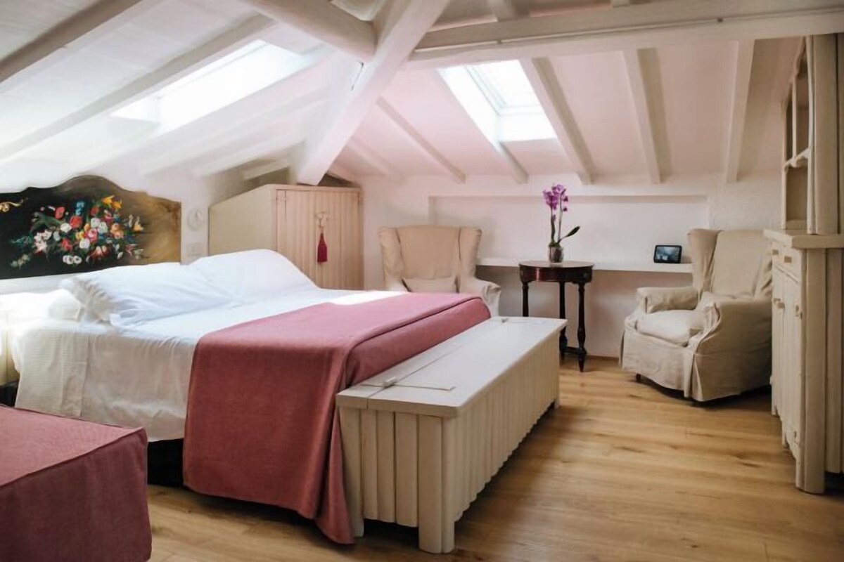 Un dormitorio con una cama, un banco, una silla, una mesita y un cuadro en la pared.