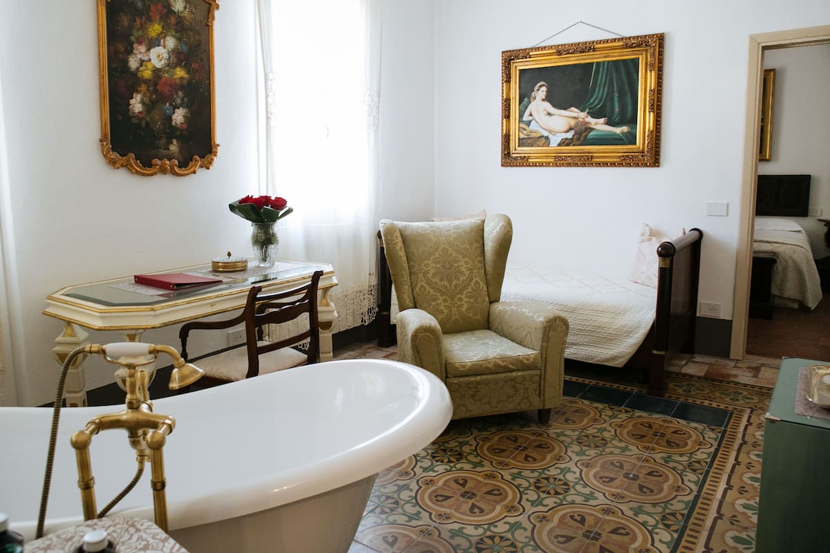 Um banheiro com banheira, uma cadeira, uma mesa com um vaso de flores e um quadro na parede.