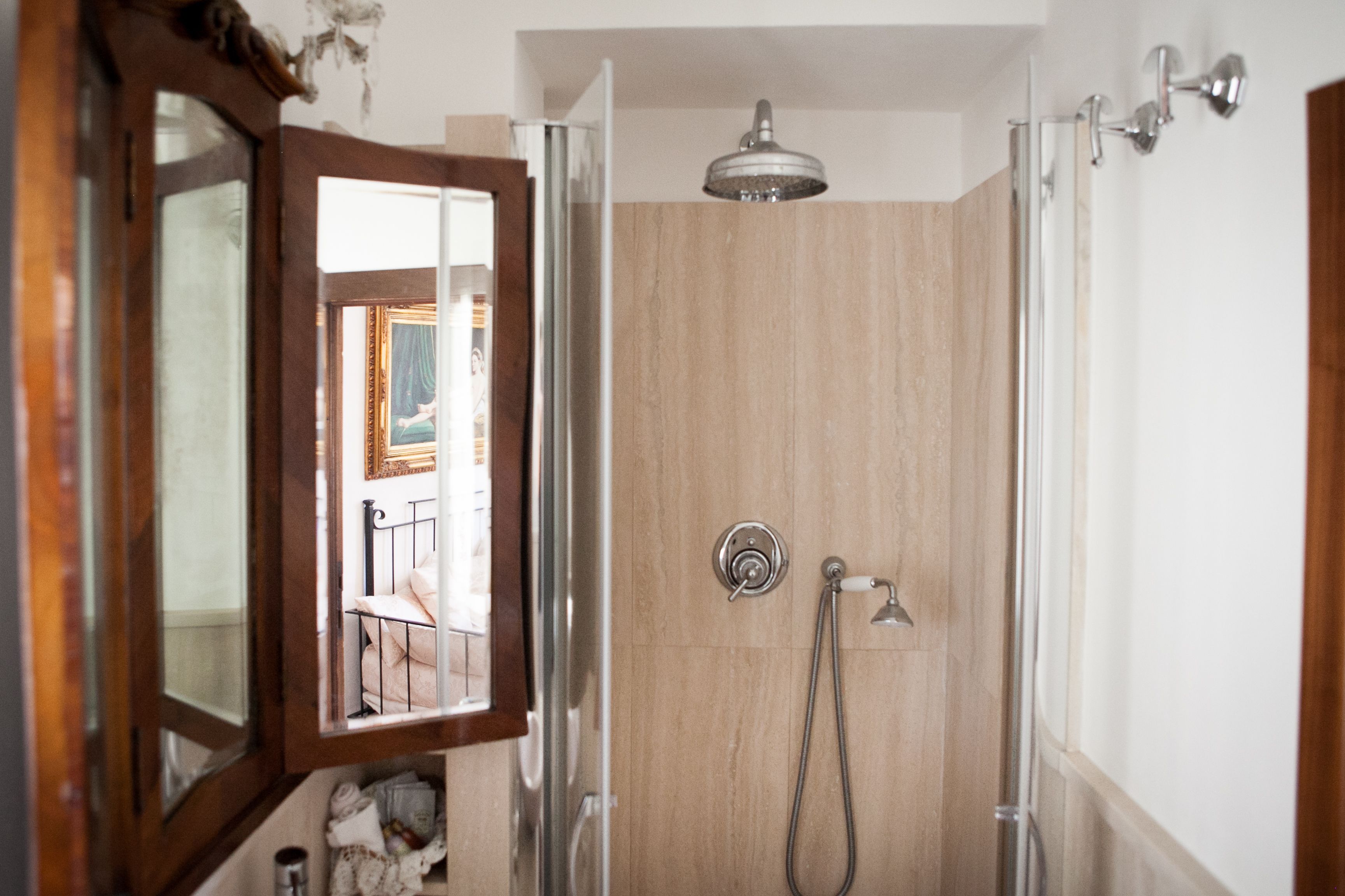 A bathroom with a shower, a mirror, and a door leading to a room with a bed and a chair.