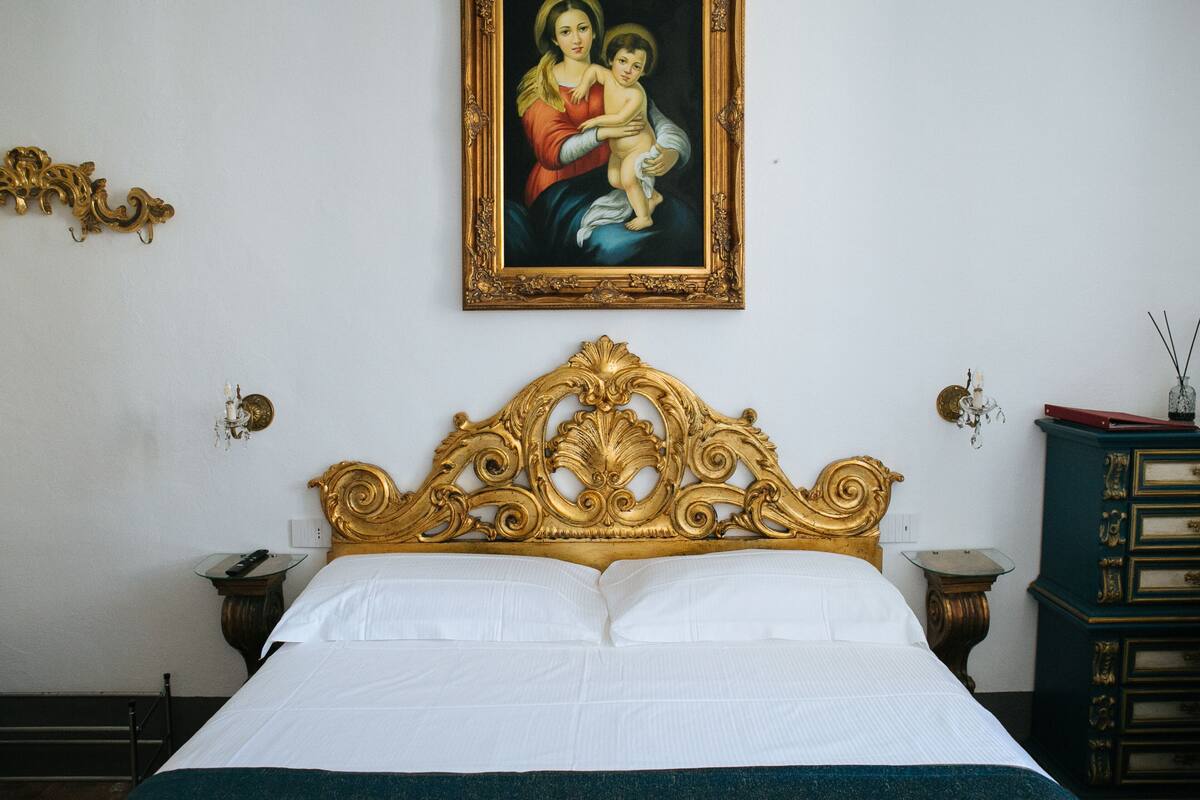 Ein Hotelzimmer mit einem großen Bett, einem verzierten Kopfteil, einem gerahmten religiösen Gemälde und einem Nachttisch mit Lampe.