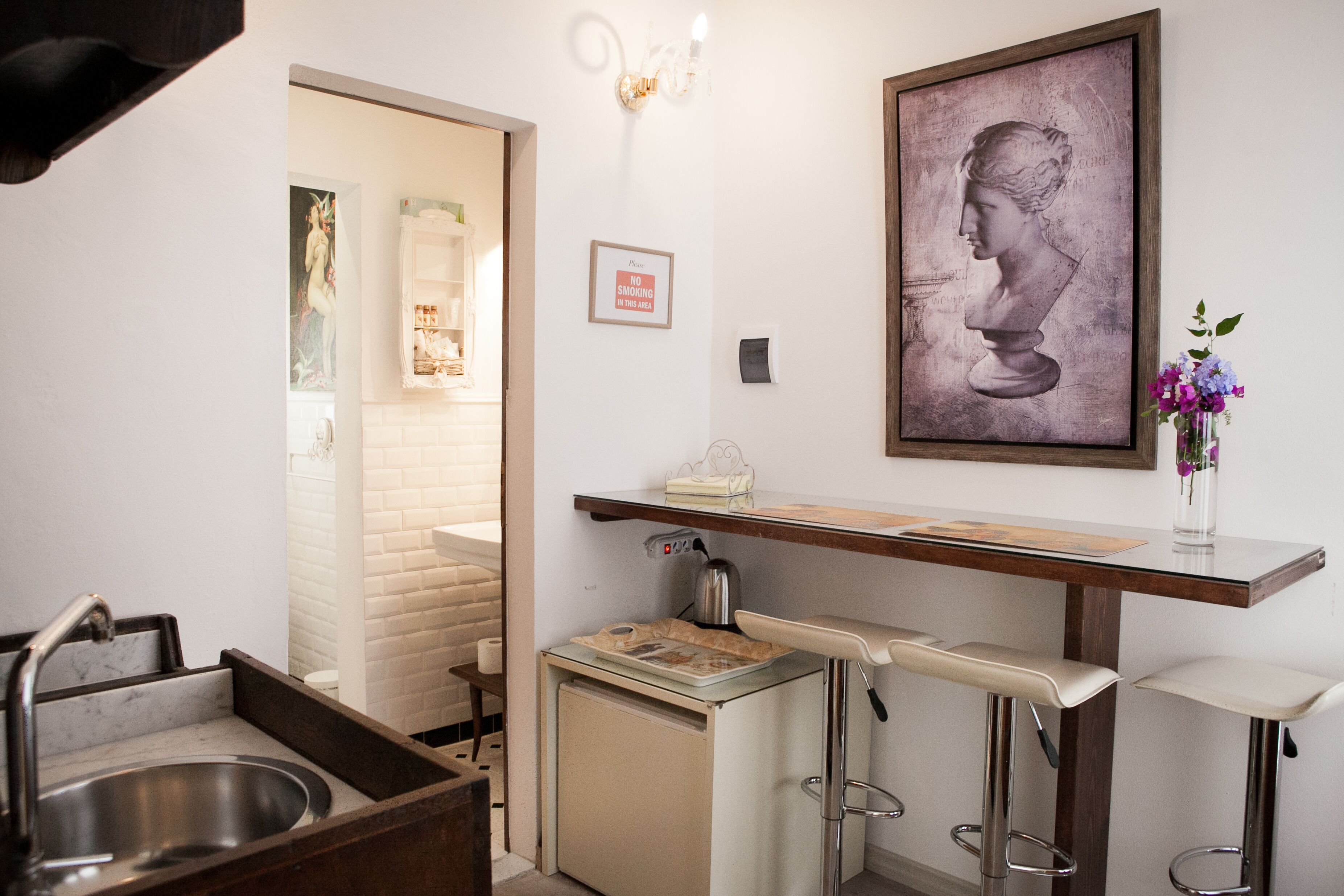 A compact kitchen with a sink, a small refrigerator, a bar stool, and a wall-mounted shelf with a framed artwork.