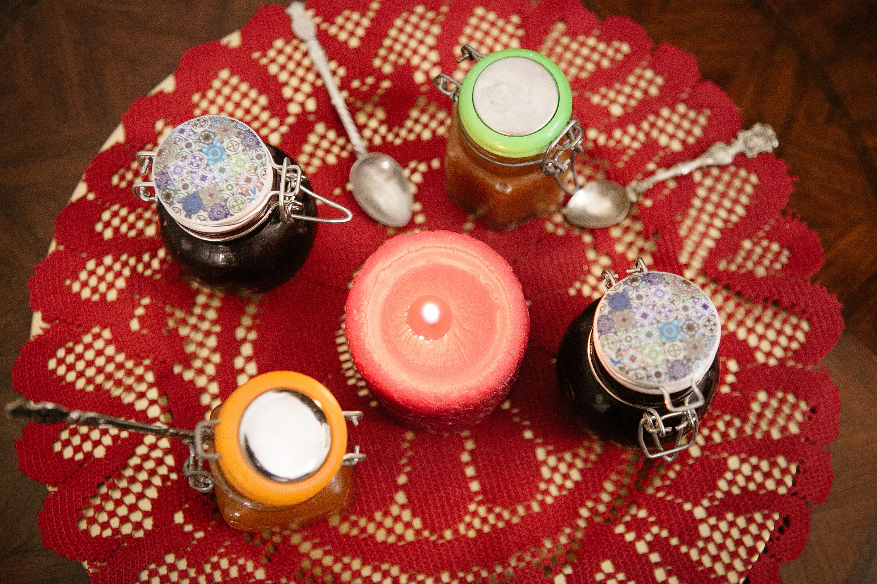 Four jars with lids, a candle, and spoons on a red crocheted doily.