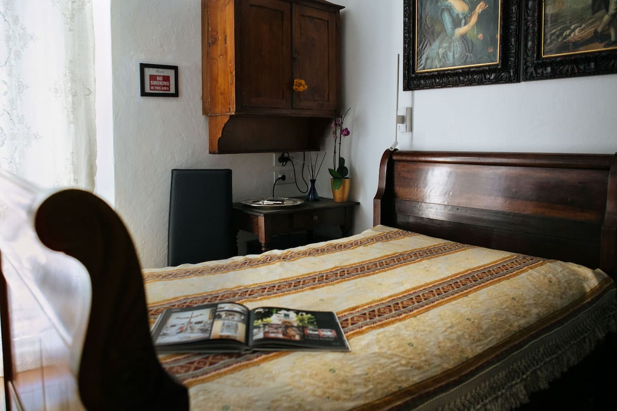 Uma cama bem arrumada com colcha estampada, cabeceira de madeira e uma pequena mesa de cabeceira com um vaso de flores.