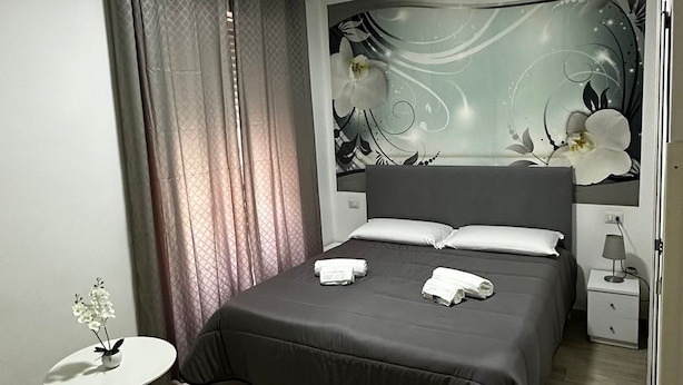 A modern bedroom with a large bed, a small bedside table, a nightstand, and a decorative wall piece. - Napoli Relais (Naples)