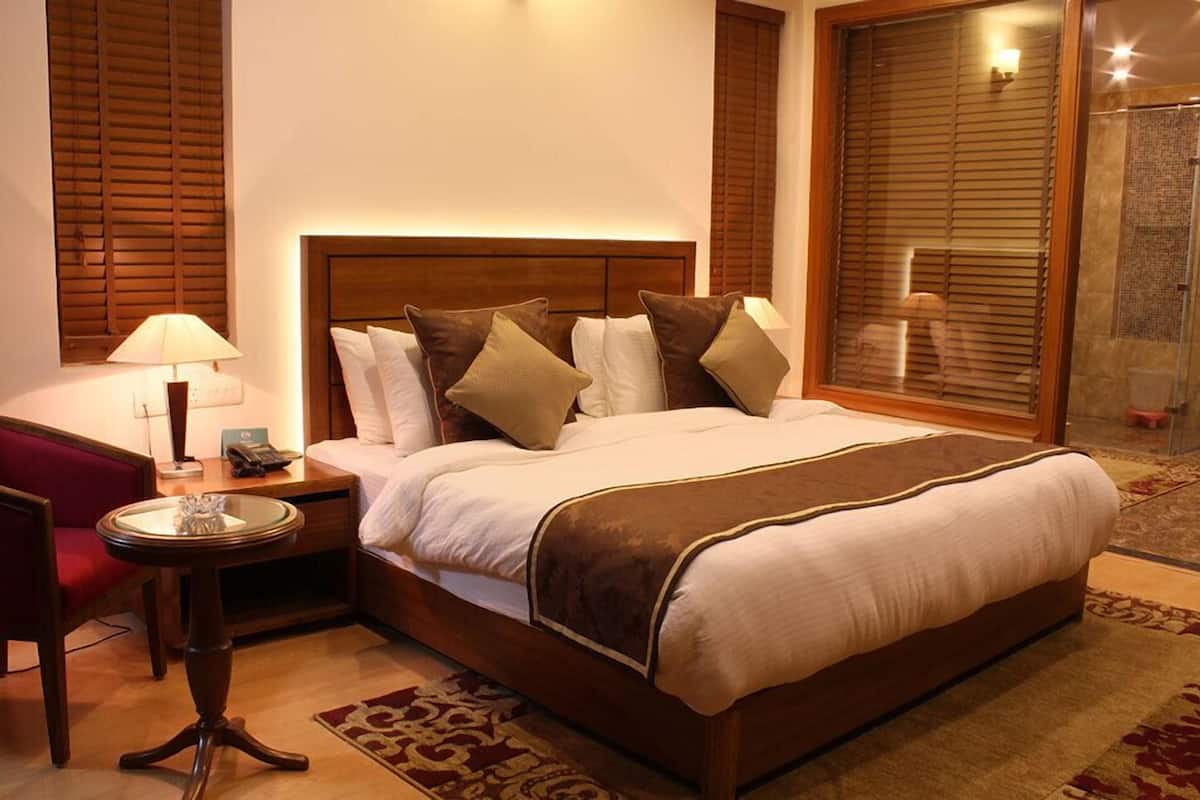 A neatly arranged hotel room with a large bed, a bedside table with a lamp, a red chair, and a window with blinds.