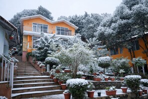 A multi-story building with a staircase leading to the entrance, surrounded by snow-covered potted plants and trees. - Essence of Nature Resort N Spa (Bhikyasain)