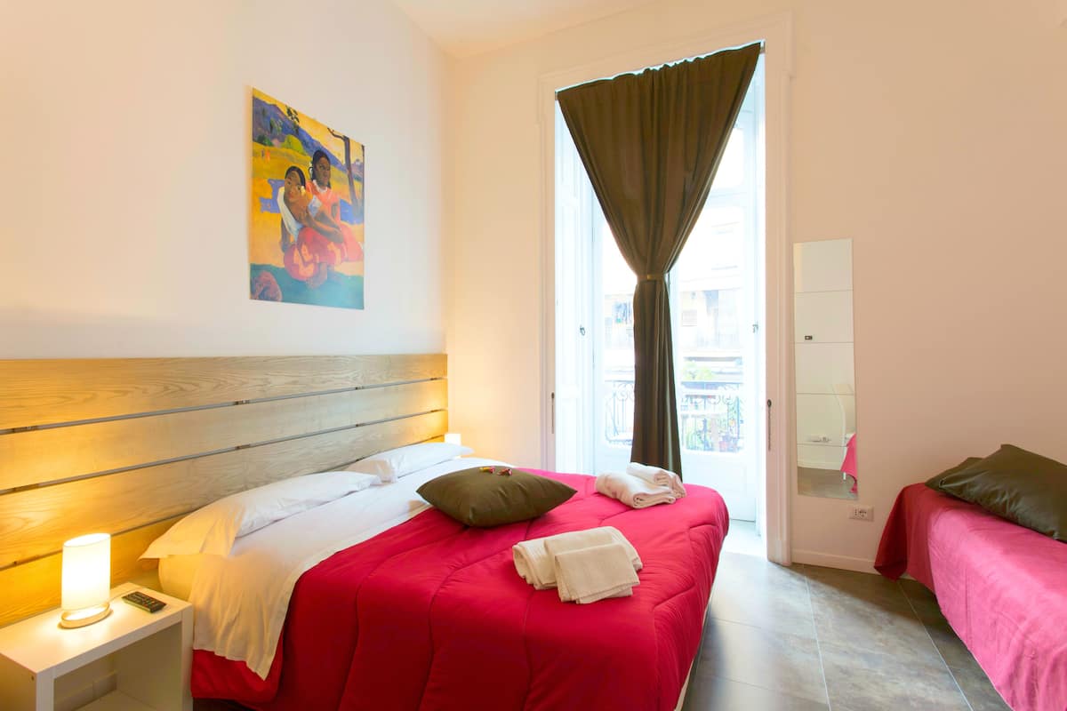 Habitación de hotel con cabecera de madera, colcha roja, almohada verde y mesita de noche blanca con lámpara.