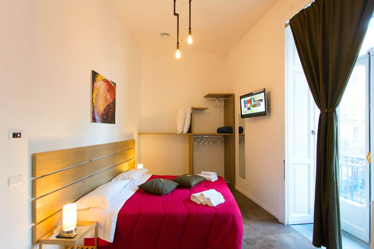 Habitación de hotel con cama, mesita de noche con lámpara, armario y ventana con cortinas.