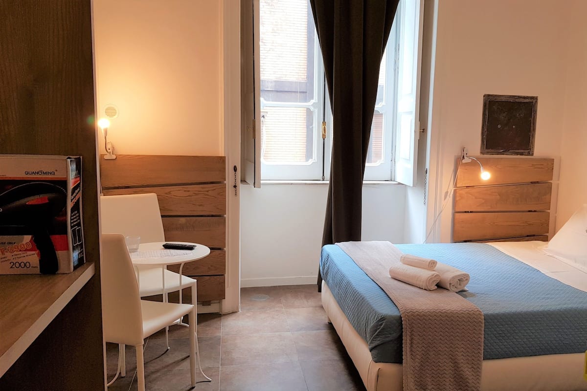 Una habitación de hotel con una cama, un escritorio con silla, una mesita, una ventana con cortinas y una mesita de noche con lámpara.