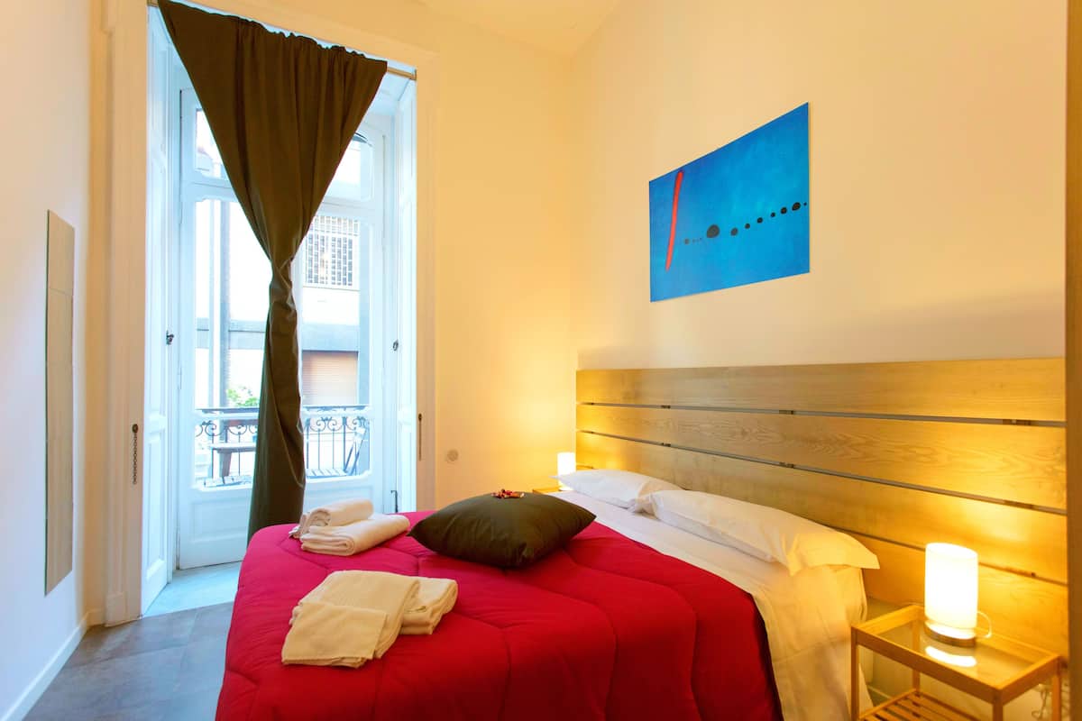 Una habitación de hotel con una cama grande, un cabecero de madera, una mesita de noche con lámpara, vistas al balcón y una obra de arte azul en la pared.