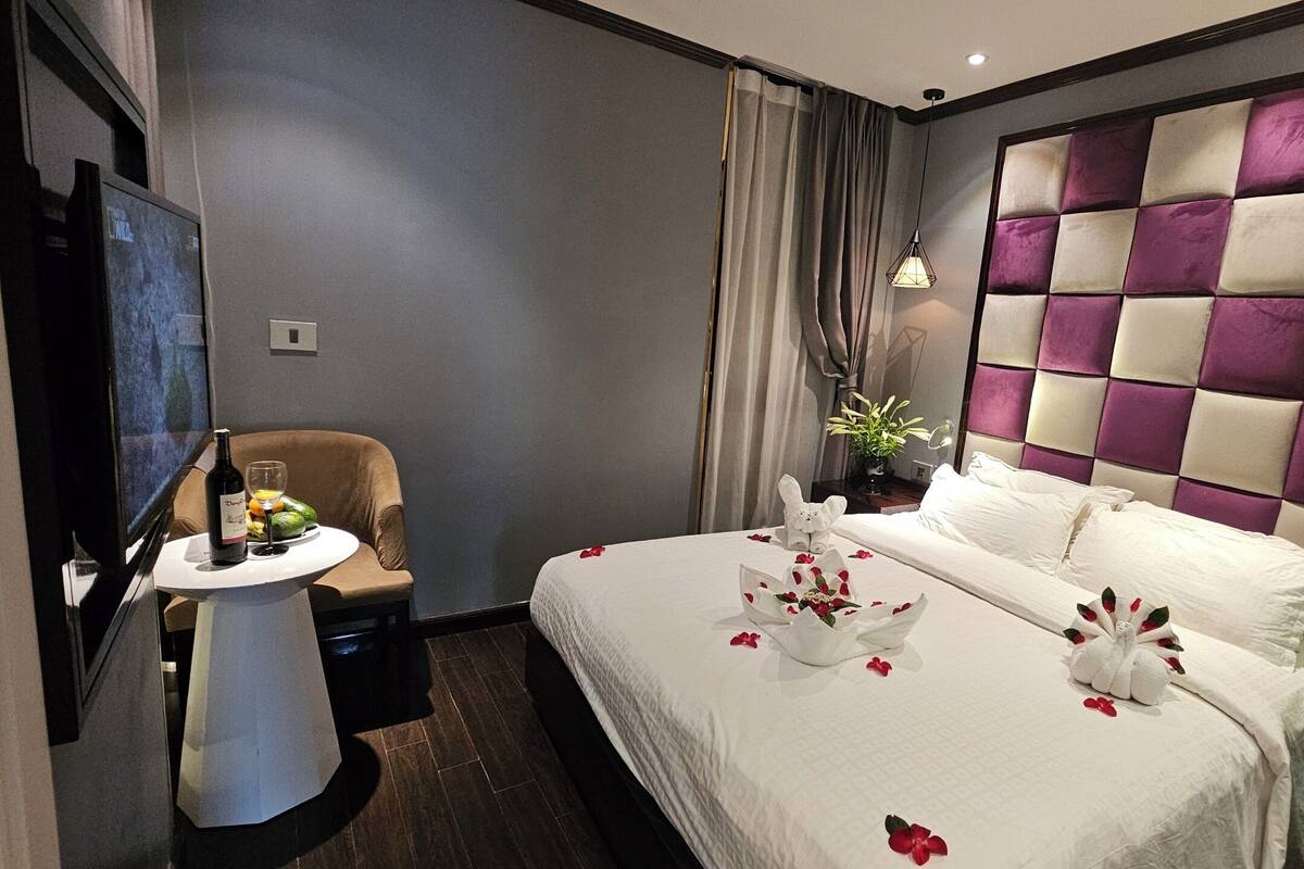 A hotel room with a bed, a small table with a bottle and glasses, a chair, and a TV on the wall.