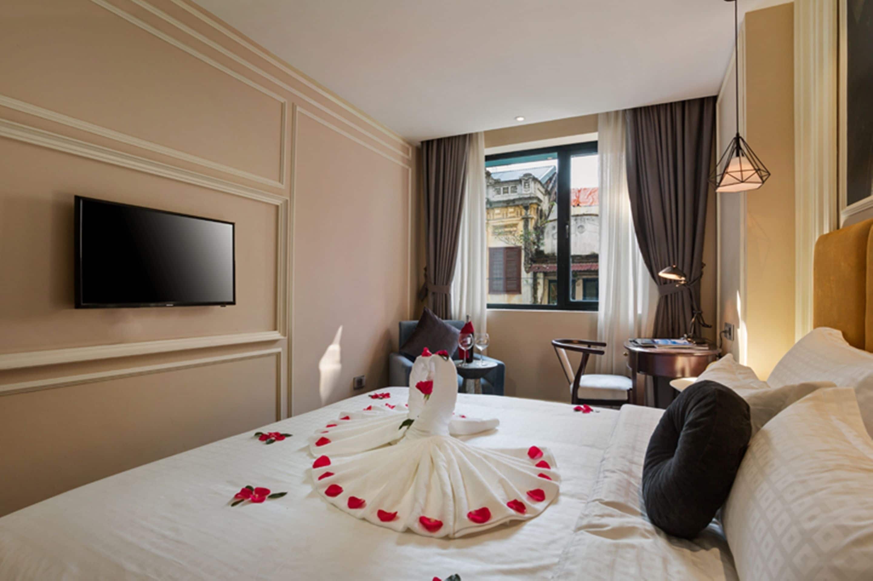 A hotel room with a bed, a flat-screen TV mounted on the wall, a small table with a lamp, and a view of the city through the window.