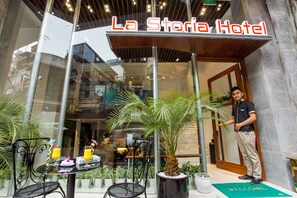 The entrance of La Storia Hotel with a glass facade, indoor plants, and a welcome mat. - Hanoi La Storia Hotel (Hanoi)