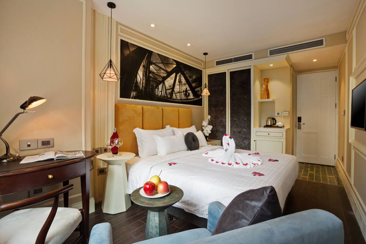 A hotel room with a bed, a desk, a small table with fruit, a chair, a TV, and a painting on the wall.