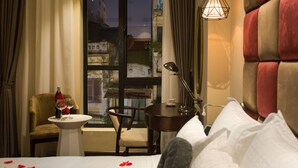 Hotelkamer met een bed, een klein tafeltje met een wijnglas en een kopje, een stoel, en een uitzicht op een stadsbeeld door een raam.
