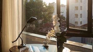 Een tafel met een lamp, een kopje en een vaas met bloemen, bij een raam met uitzicht op gebouwen.