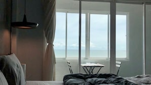 A bedroom with a bed, a small table, and a chair. It has a large window with a view of the sea. - Betta Hostel (Chonburi)