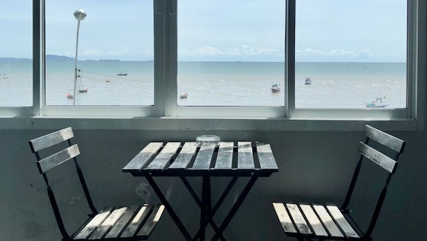 A view from a window with a table and two chairs, overlooking a calm sea with boats and a distant shoreline. - Betta Hostel (Chonburi)