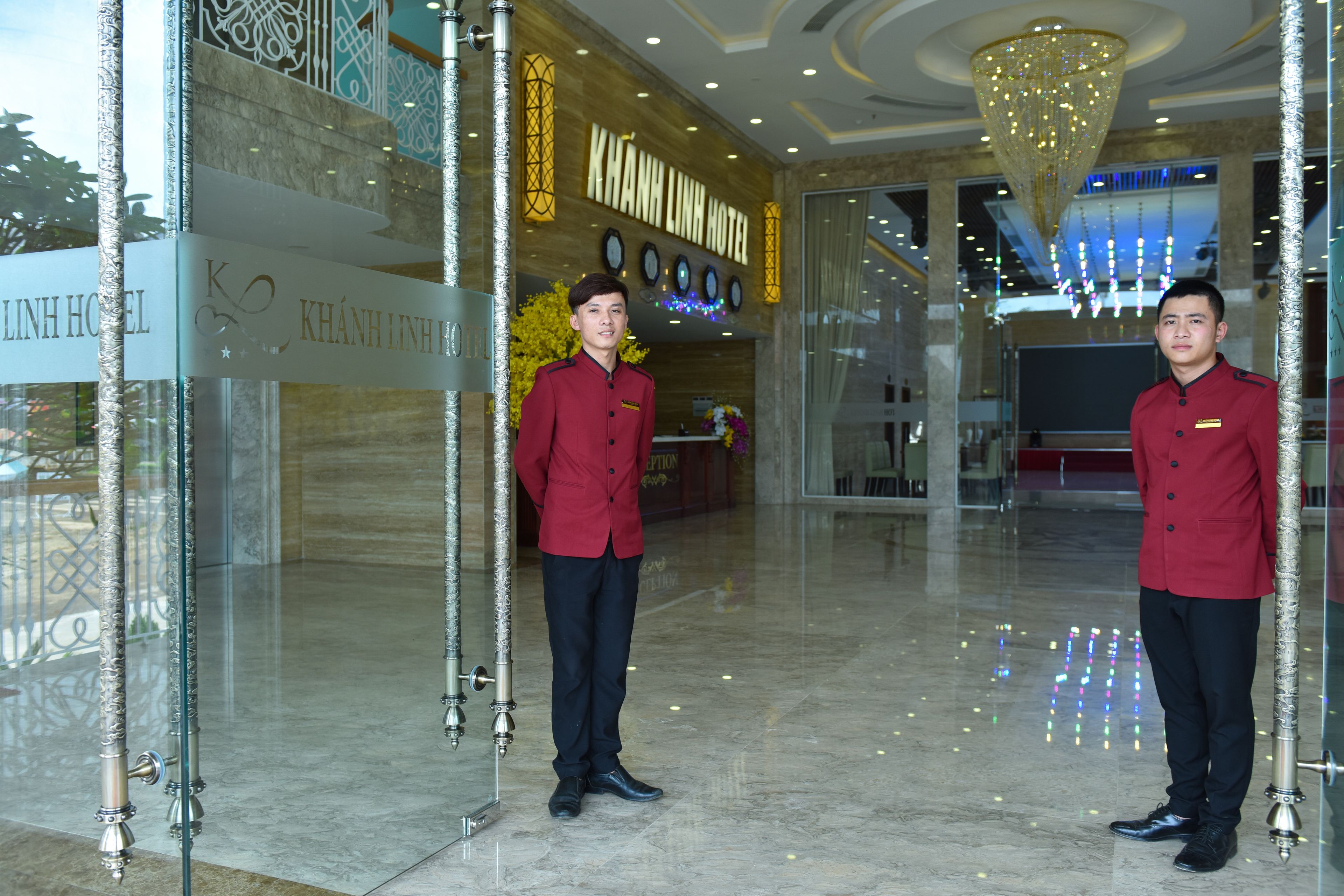 A hotel lobby with a reception desk, a chandelier, and two uniformed staff members.