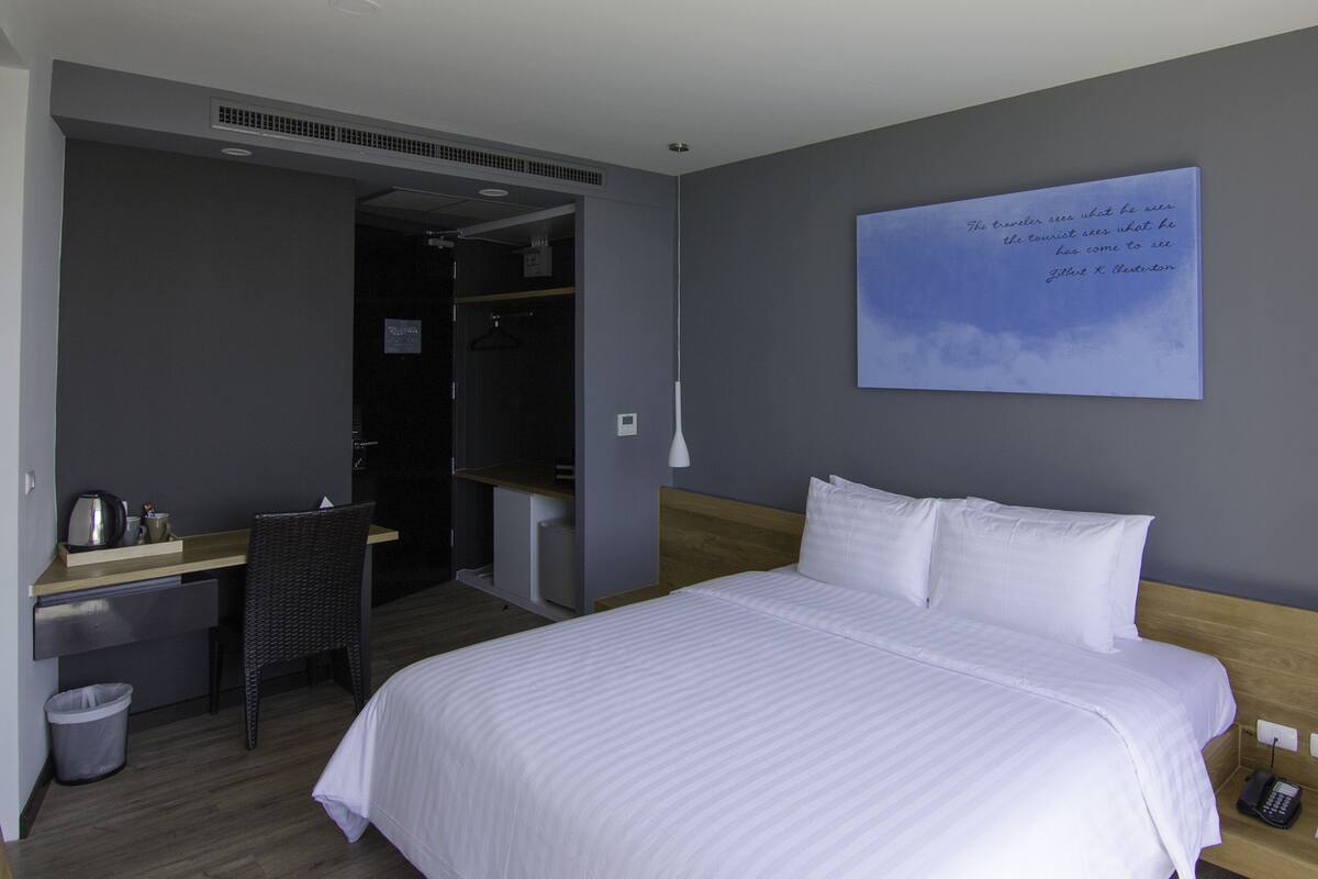 A hotel room with a bed, a desk, a chair, a waste bin, and a wall-mounted artwork.