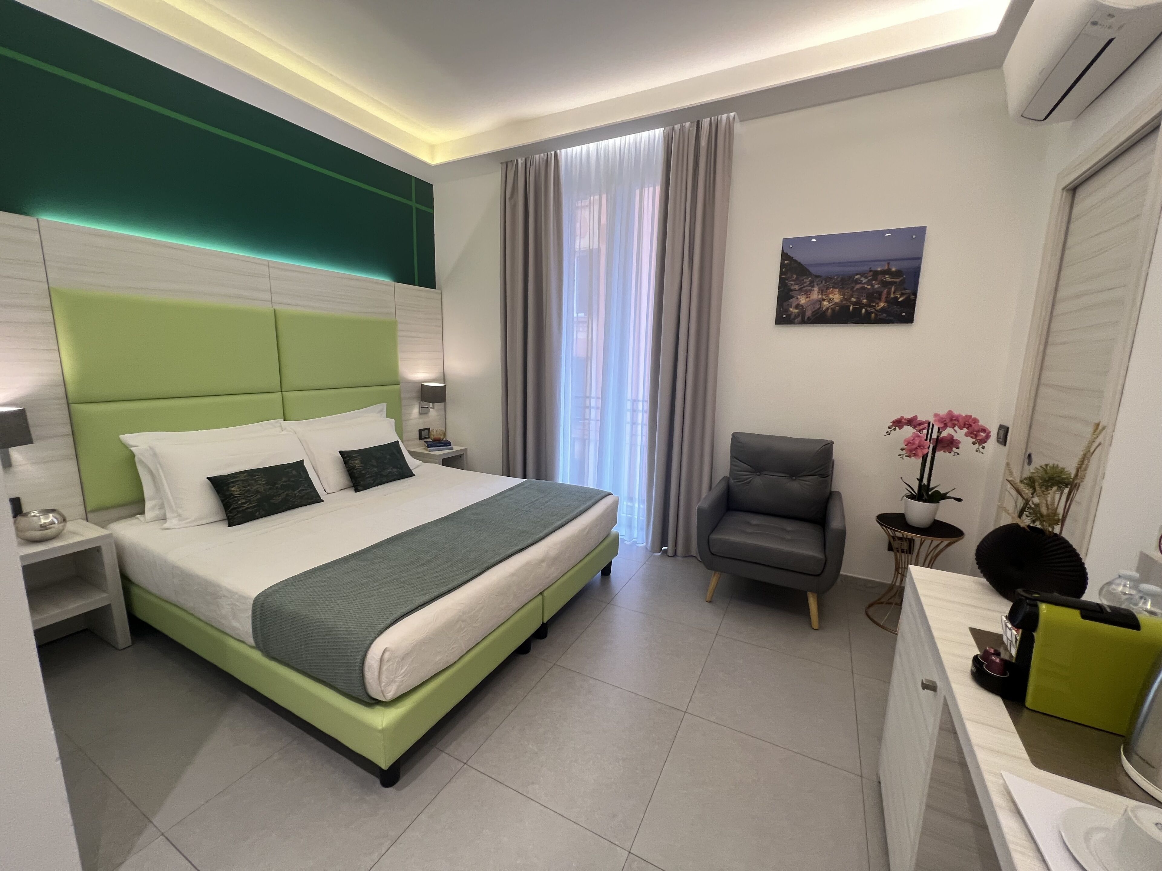 Una habitación de hotel moderna con una paleta de colores verde y blanco, una cama grande, un sillón gris, una mesita con un jarrón de flores y un cuadro con una vista urbana en la pared.