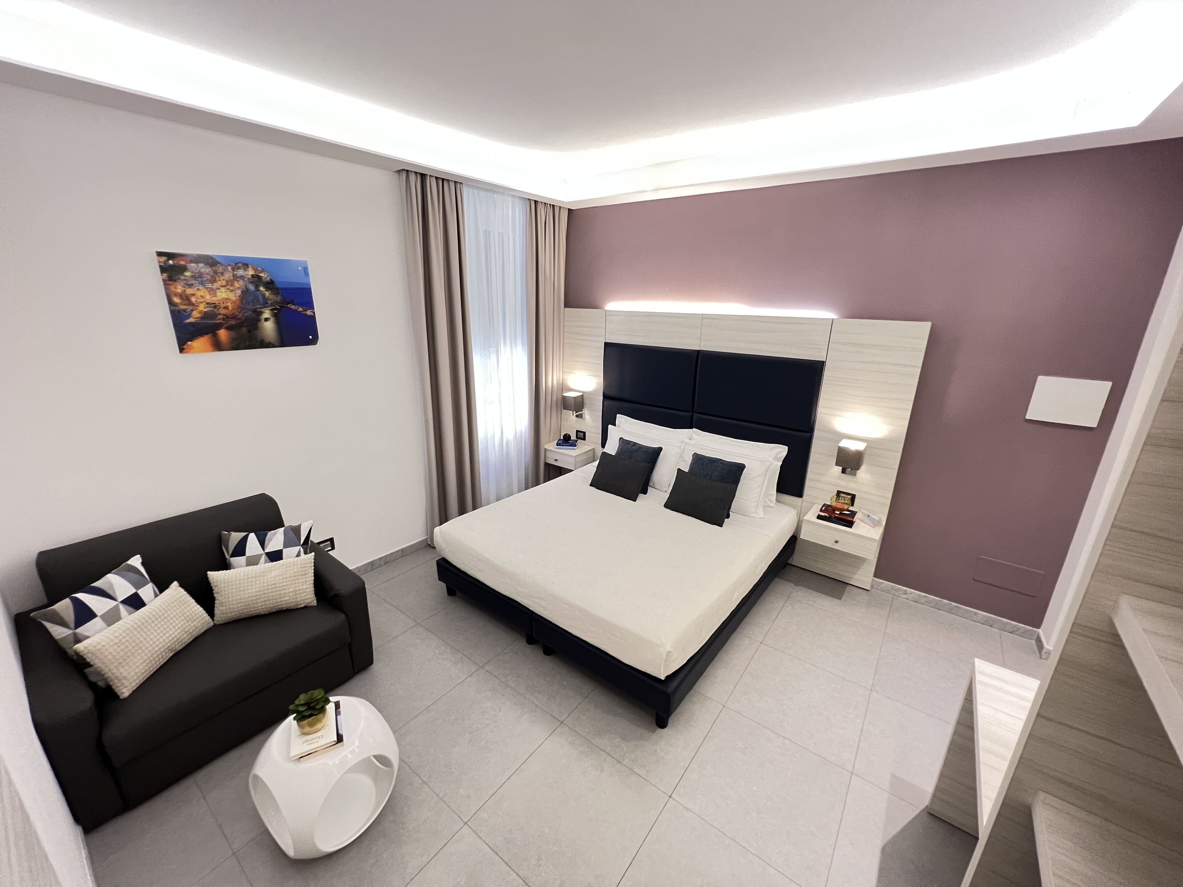 A modern hotel room with a large bed, a black sofa, a small round table, and a painting on the wall.