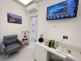 Una habitación de hotel moderna con un televisor de pantalla plana mostrando un noticiero, un sofá gris cómodo, una pequeña mesita de noche con un jarrón de flores rosadas y una zona de kitchenette con cafetera y tetera.