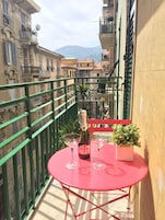 Una mesa roja con una botella de Valmarone Secco y dos copas en un balcón con barandilla verde, con vistas a un paisaje urbano que incluye edificios y montañas.