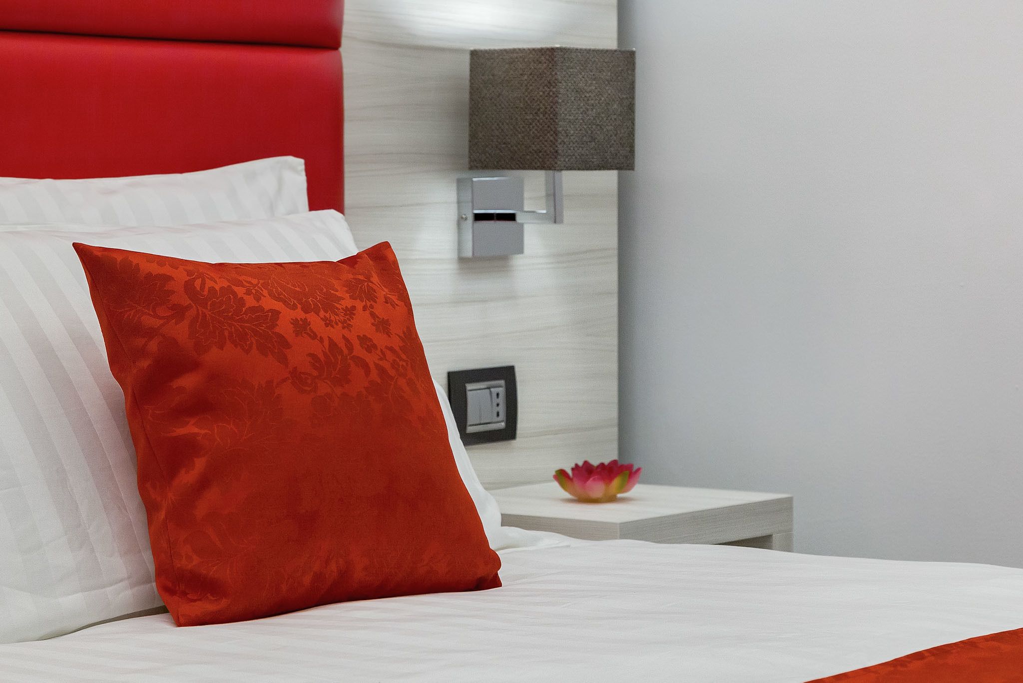 A bed with white and red pillows, a red headboard, a nightstand with a lamp, and a decorative bowl.