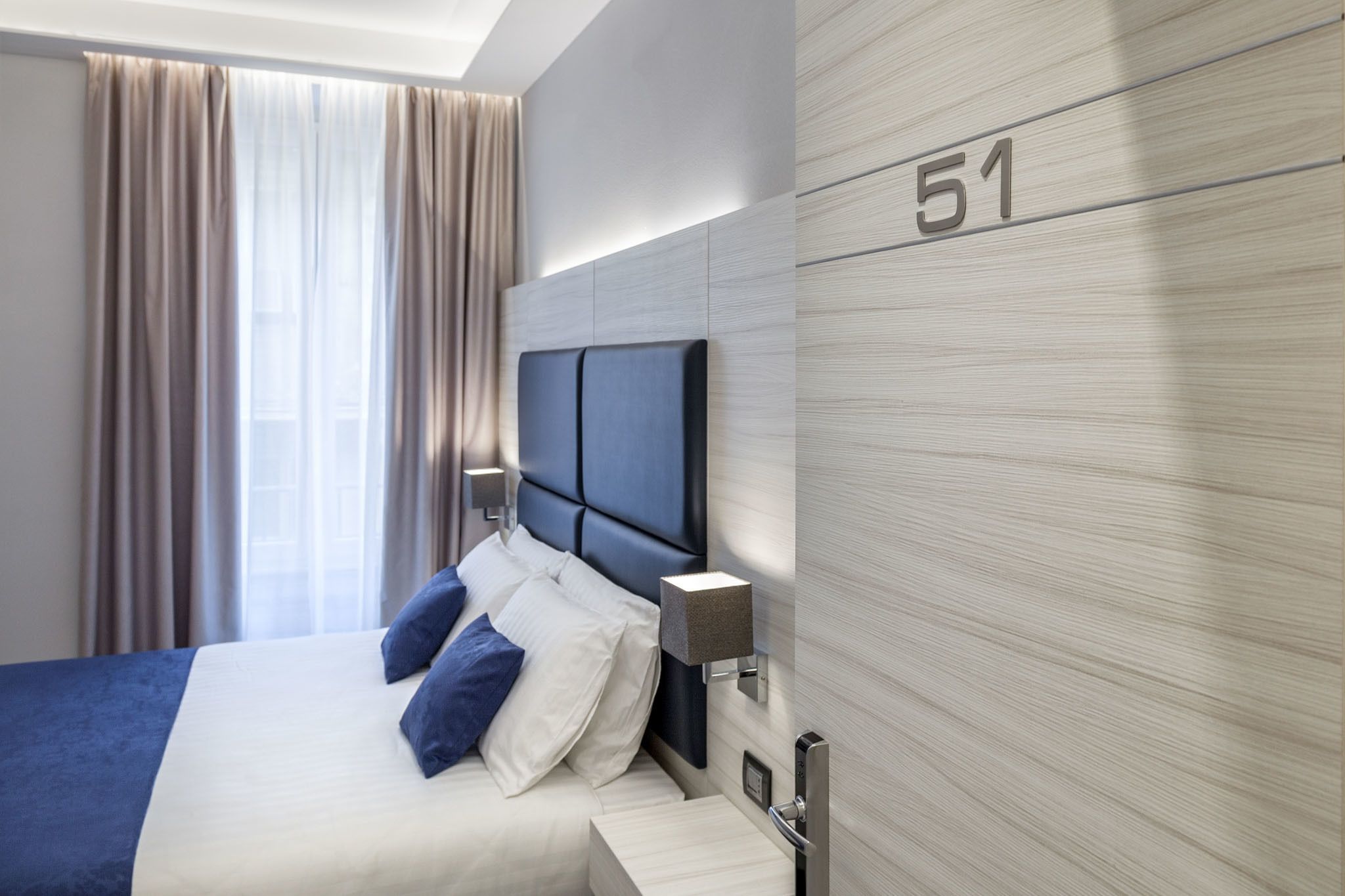 A modern hotel room with a large bed, a headboard, a bedside table, a lamp, and a wall-mounted number '51'.