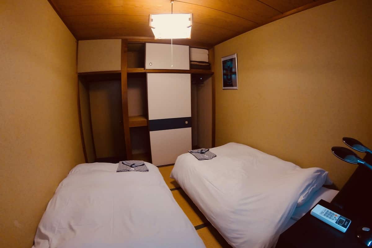 A small hotel room with two single beds, a wardrobe, a desk with a computer, and a nightstand with a lamp.