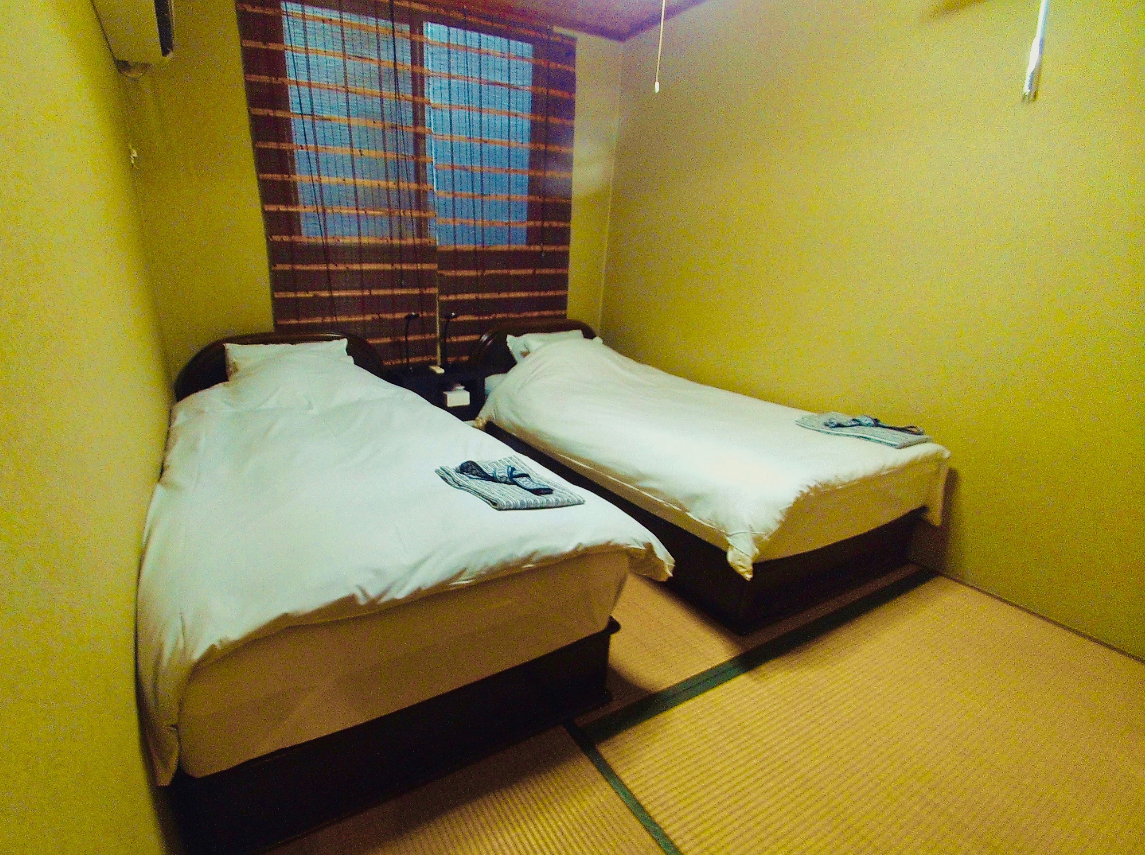 Ein kleines Zimmer mit zwei Betten, einem Fenster mit Jalousien und einer Fußmatte.