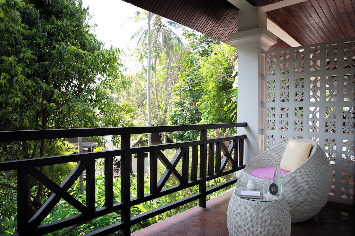 A balcony with a wicker chair and a small table, surrounded by lush greenery and a white lattice fence.