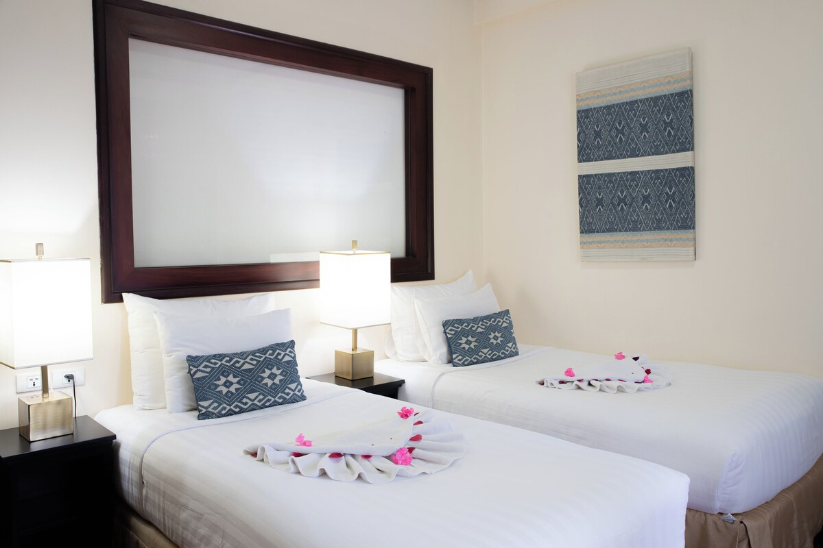 Quarto de hotel com duas camas, um espelho grande, uma obra de arte na parede e uma mesa de cabeceira com abajur.