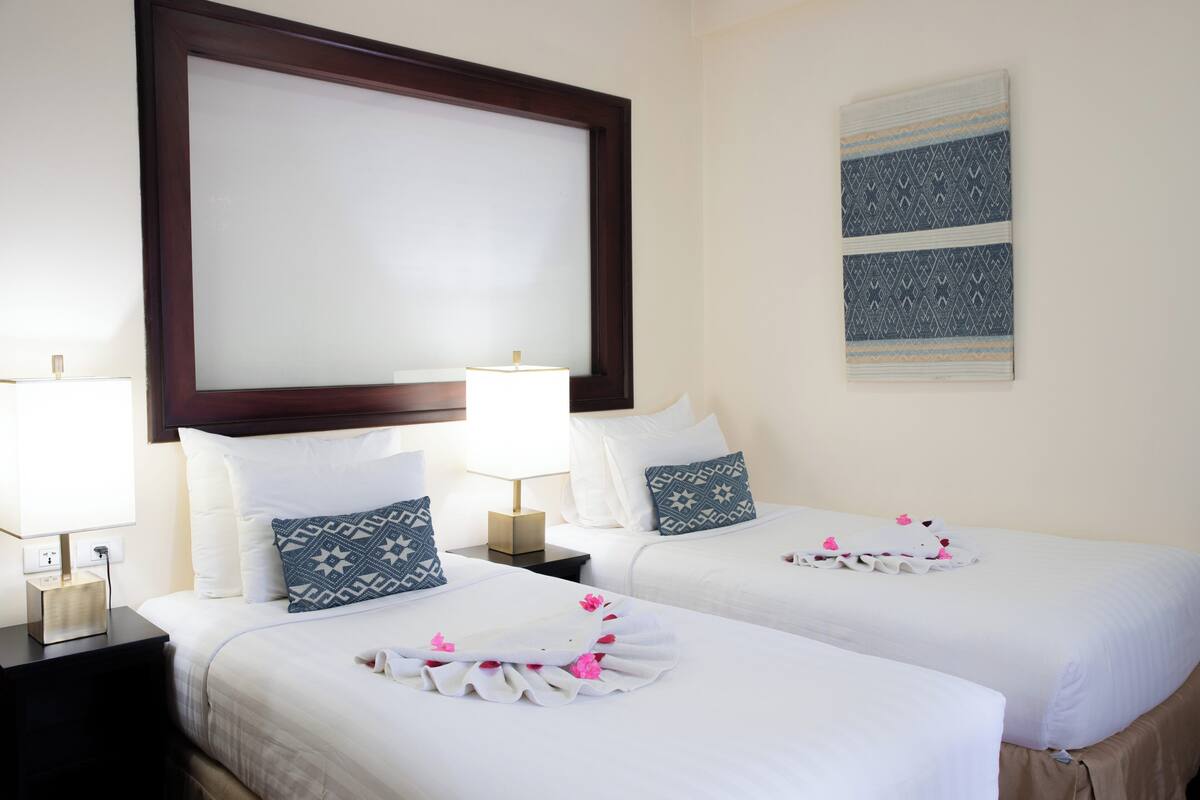 A hotel room with two beds, a large mirror, a wall-mounted artwork, and a bedside table with a lamp.