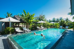 A swimming pool with two people swimming, surrounded by lounge chairs and umbrellas, with palm trees and a clear blue sky in the background. - Luna Villa Homestay (Da Nang)