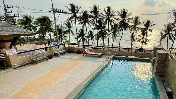 A rectangular swimming pool with clear blue water, surrounded by a tiled deck area with benches and a small structure. Palm trees are visible in the background, suggesting a tropical setting. - La Playa Boutique Resort (Chonburi)