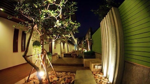 A nighttime outdoor area with a wooden deck, a tree, and a green building. - La Playa Boutique Resort (Chonburi)