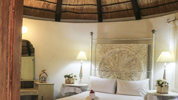 A bedroom with a thatched roof, a bed with white linens, two bedside tables with lamps, and a nightstand with a clock. - The Green Acorn Guest House (Setsoto)