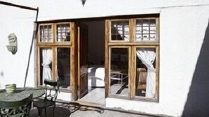 A white building with a wooden door and windows, a small table with chairs, and a lamp on the wall. - The Green Acorn Guest House (Setsoto)